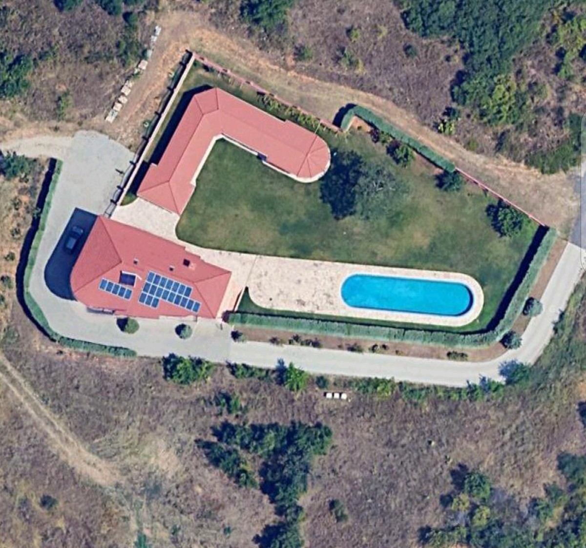На сателитната снимка от птичи поглед се разкрива мащабът на едно истинско феодално имение, чиято конфигурация поражда сериозни въпроси относно произхода на средствата за неговото изграждане. В центъра на парцела доминира масивна жилищна сграда с Г-образна форма и червен скатен покрив, върху който ясно се виждат инсталирани множество соларни панели — детайл, подчертаващ стремежа към енергийна независимост на имота. Най-впечатляващият елемент обаче е огромният басейн с яркосиня вода, заобиколен от обширна, професионално поддържана тревна площ и прецизно оформена декоративна растителност по периферията, която служи за естествена преграда от любопитни погледи. Цялото имение е опасано от солидна ограда и асфалтиран вътрешен път, който води до закрити гаражи, а общата площ на облагородения терен рязко контрастира с необработените и пусти съседни парцели, визуално потвърждавайки хипотезата на Топ Преса за натрупване на значително богатство, инвестирано в лукс, който трудно може да остане незабелязан от данъчните власти.